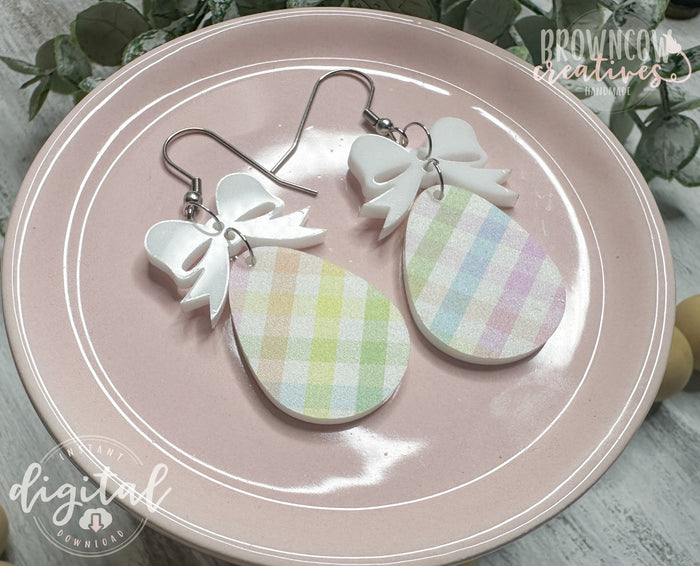 A pair of handmade Easter-themed earrings displayed on a pink plate. The earrings feature a pastel plaid teardrop-shaped charm hanging from a white bow-shaped topper, attached with metal hooks. The background includes greenery and branding for "Brown Cow Creatives." A watermark indicates that this is a digital download for a laser-cut file.