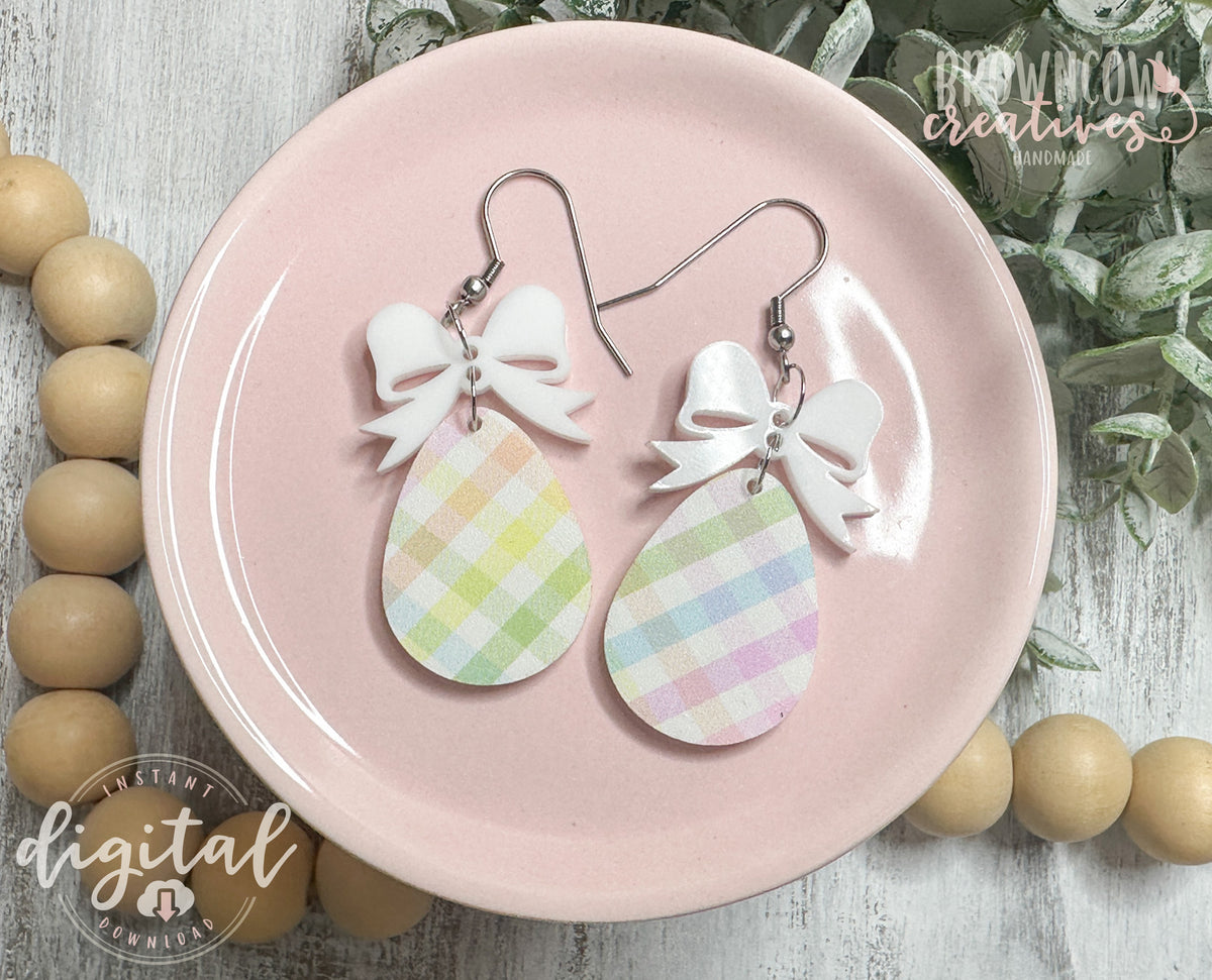 A pair of handmade Easter-themed earrings displayed on a pink plate. The earrings feature a pastel plaid teardrop-shaped charm hanging from a white bow-shaped topper, attached with metal hooks. The background includes greenery and branding for "Brown Cow Creatives." A watermark indicates that this is a digital download for a laser-cut file.