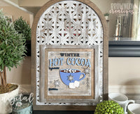 A decorative laser-cut Winter Hot Cocoa Sign displayed in a rustic wooden and metal arched frame. The sign features layered text and engraved details, including a blue mug filled with marshmallows, snowflakes, and recipe instructions for hot cocoa. The background has a distressed wood aesthetic, enhancing the cozy winter theme. Surrounding the sign are home decor elements such as potted greenery, a candle, and white mugs. 